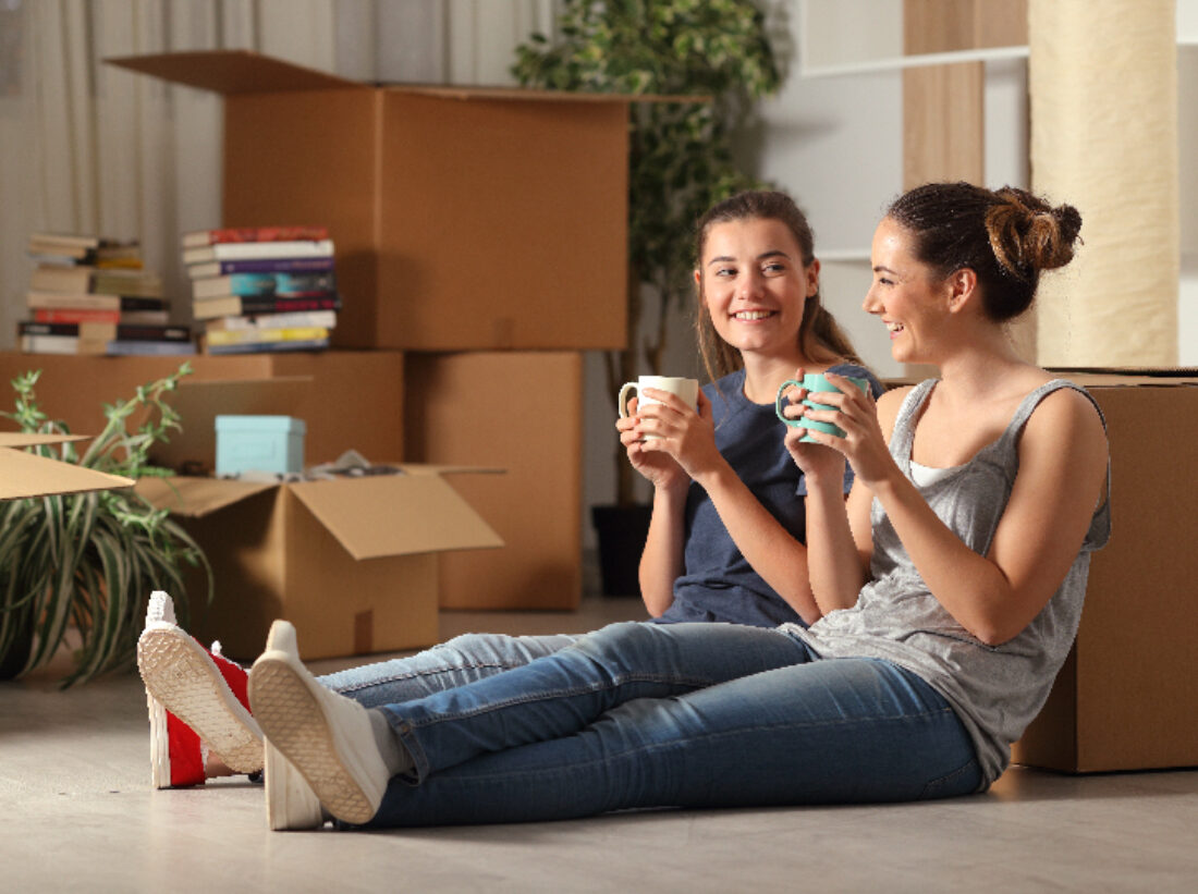two women sitting on the floor with moving boxes around them, catching up with a cup of tea
