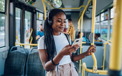 Young black woman wearing earphones and listening to music on a smartphone while standing alone on a bus.