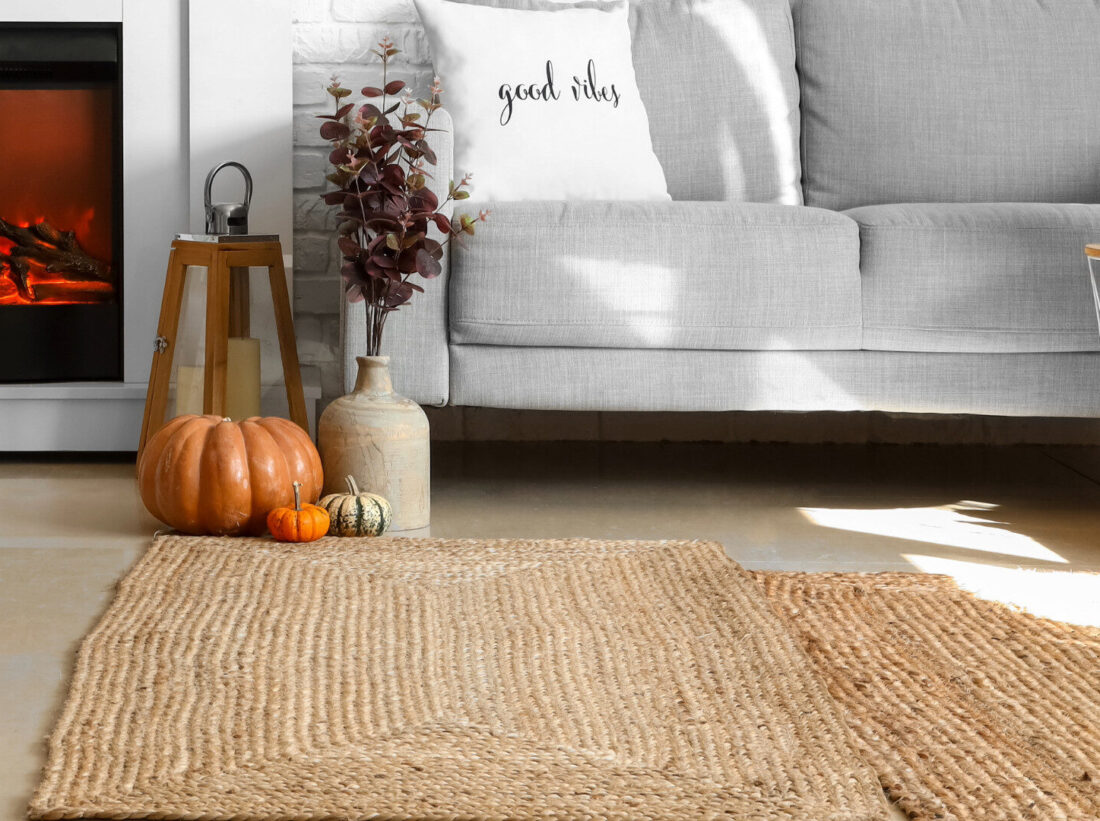 A textured straw rug on a laminate flooring in a living room, in front of a white sofa with a throw cushion, surrounded by autumnal pumpkin decorations