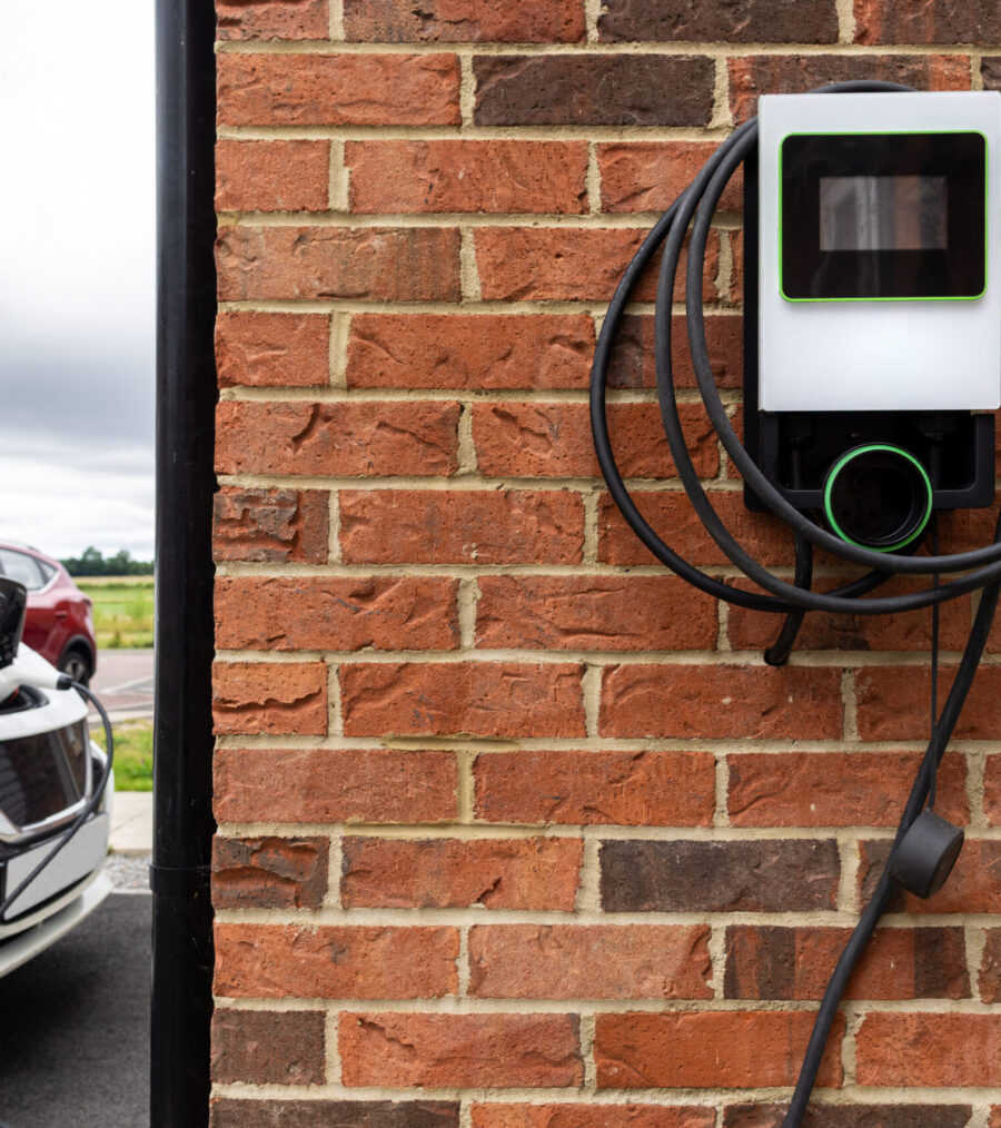 A medium shot of a white electric vehicle parked in a residential driveway while charging from a wall-mounted electric car charger attached to a red brick house. The charging cable is connected to the car’s front port, representing sustainable energy, eco-friendly transportation, and a modern suburban lifestyle.